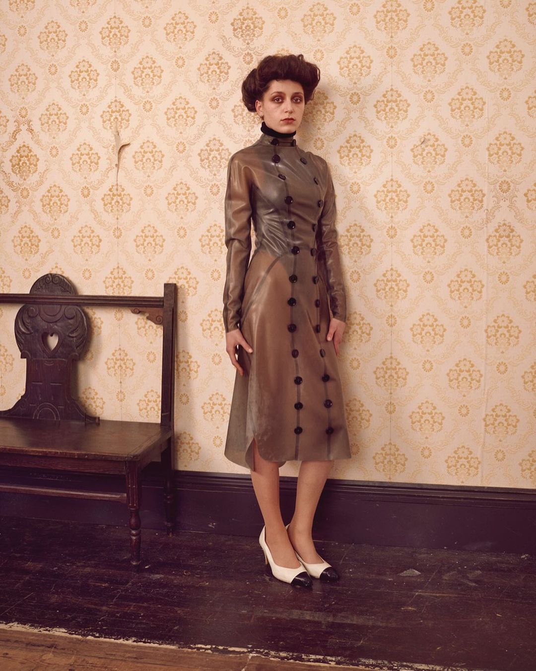 Woman in a vintage-inspired translucent dress and two-tone heels poses against patterned wallpaper, standing near a vintage wooden bench.