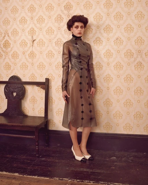 Woman in a vintage-inspired translucent dress and two-tone heels poses against patterned wallpaper, standing near a vintage wooden bench.