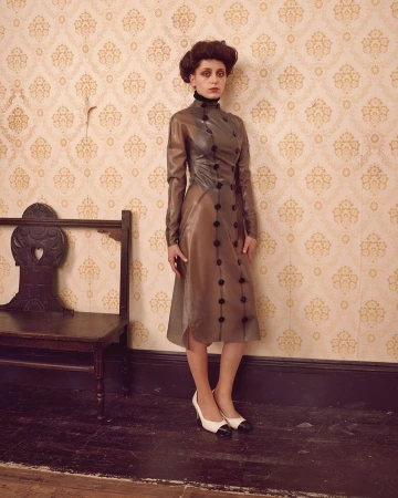Woman in a vintage-inspired translucent dress and two-tone heels poses against patterned wallpaper, standing near a vintage wooden bench.