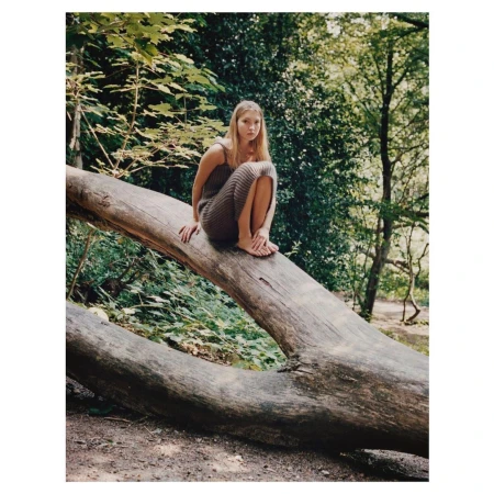 A blonde woman in a brown dress sits barefoot on a fallen tree in a green forest, gazing forward in a tranquil outdoor scene.