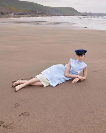 Model wearing a light blue dress and dark cap reclining barefoot on a wide sandy beach