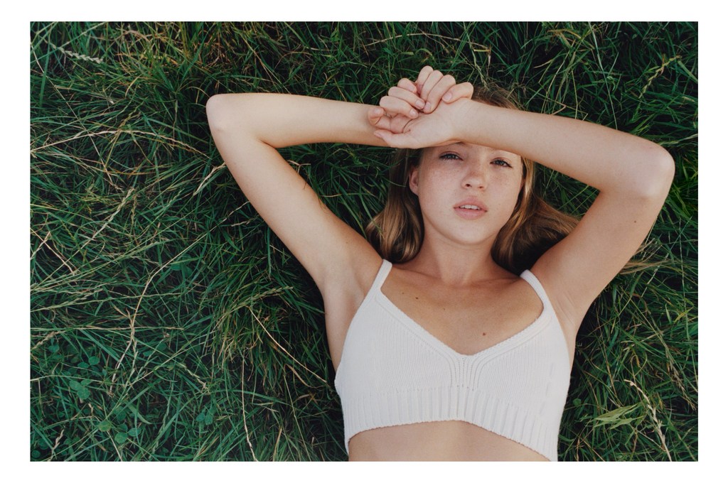 Young woman lying on green grass with arms overhead, wearing a white crop top, photographed with a soft focus and natural light.