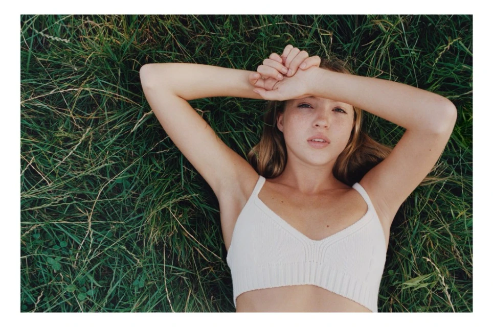Young woman lying on green grass with arms overhead, wearing a white crop top, photographed with a soft focus and natural light.