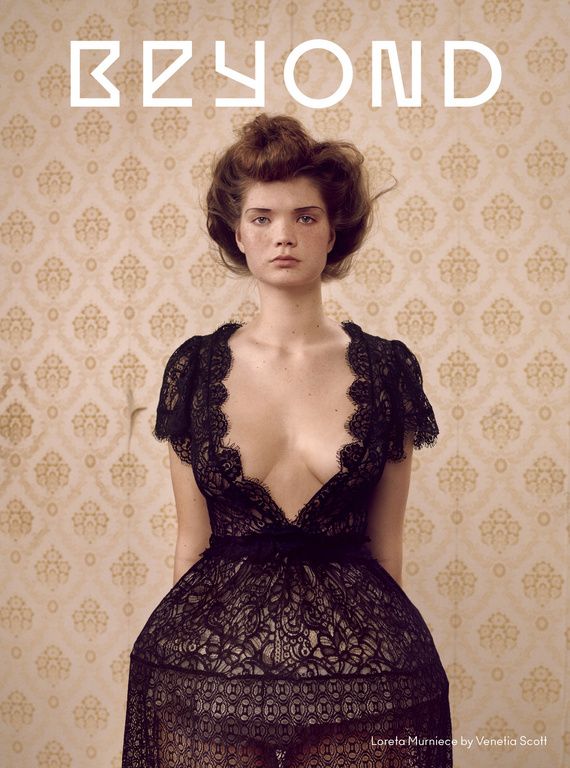 Portrait of a woman with fair skin and brown hair, wearing a black lace dress against a vintage floral wallpaper backdrop.