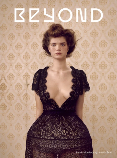 Portrait of a woman with fair skin and brown hair, wearing a black lace dress against a vintage floral wallpaper backdrop.