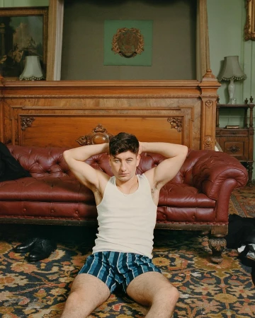A man in a white tank top and blue striped shorts seated on a patterned rug in front of a tufted leather sofa