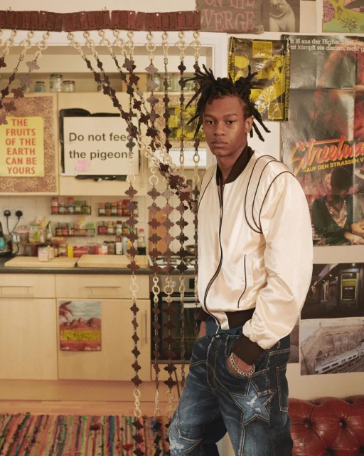 Young adult male in a cream bomber jacket and distressed jeans with posters in an indoor kitchen setting looking at the camera.