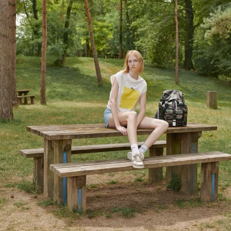 Young woman sits on a wooden picnic table in a grassy park with trees, wearing casual clothing and a backpack, in a daylight shot.
