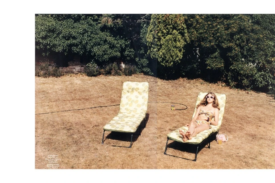 Woman in floral bikini relaxes on a patterned chaise lounge on a grassy lawn, alongside a matching chaise lounge in a backyard garden setting.