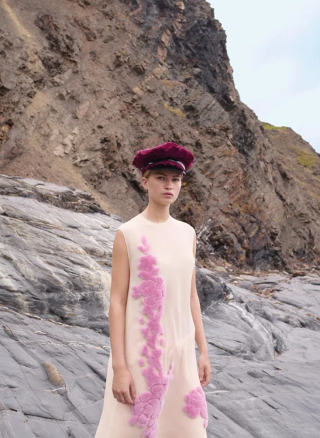 A woman in a sleeveless dress and hat stands on a rocky beach, with a large rock formation behind her in an outdoor fashion editorial.