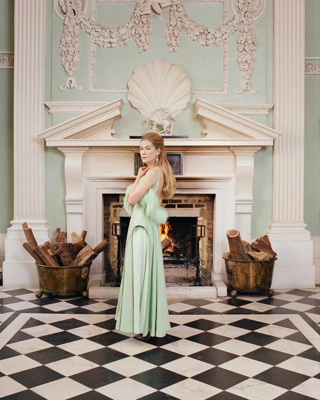 Woman in a light green gown poses in front of a fireplace with logs in a grand hall with a black and white checkered floor.