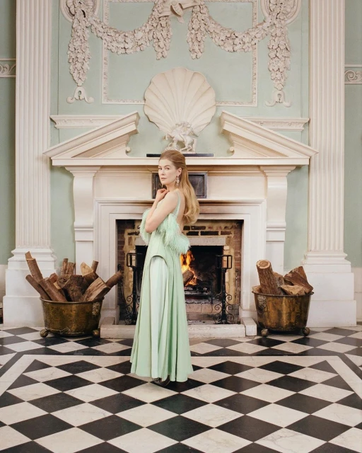 Woman in a light green gown poses in front of a fireplace with logs in a grand hall with a black and white checkered floor.