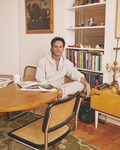 Indoor portrait of a man in pajamas seated at a round table with books, bookshelves, and a painting of a lion in the background.