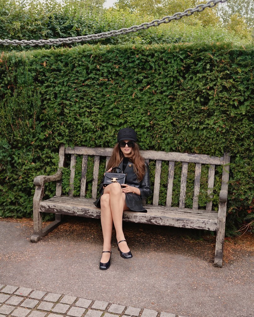 Young woman in black hat and coat sitting on a park bench, holding black purse, in front of a green hedge on a sunny day.