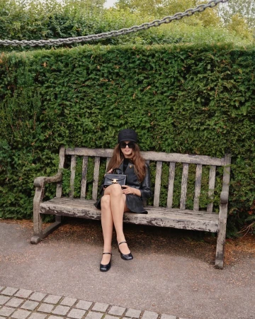 Young woman in black hat and coat sitting on a park bench, holding black purse, in front of a green hedge on a sunny day.