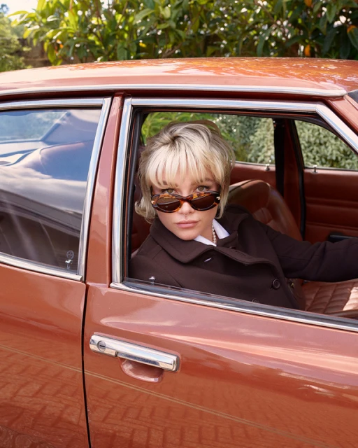 A blonde woman wearing sunglasses sits in the driver's seat of an old brown car, with green leafy trees in the background.