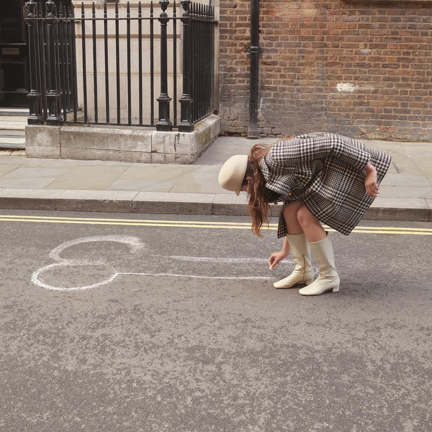 Woman in beige boots and coat drawing phallic chalk art on city street with brick buildings in the background on a sunny day.