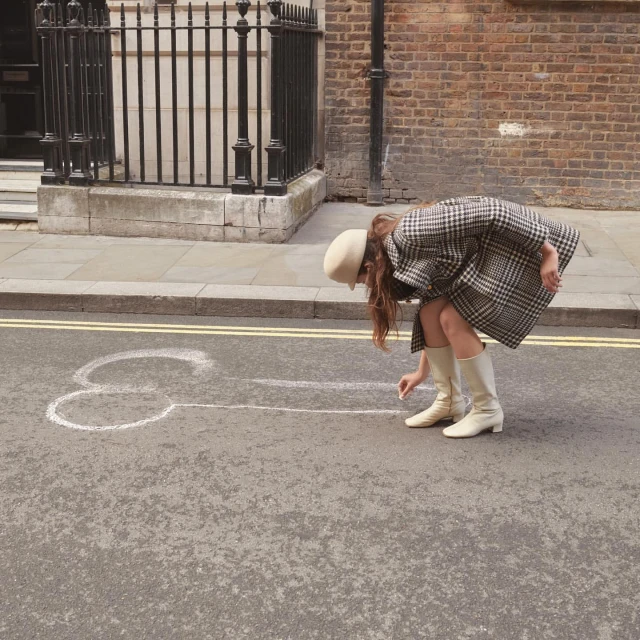 Woman in beige boots and coat drawing phallic chalk art on city street with brick buildings in the background on a sunny day.