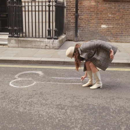 Woman in beige boots and coat drawing phallic chalk art on city street with brick buildings in the background on a sunny day.