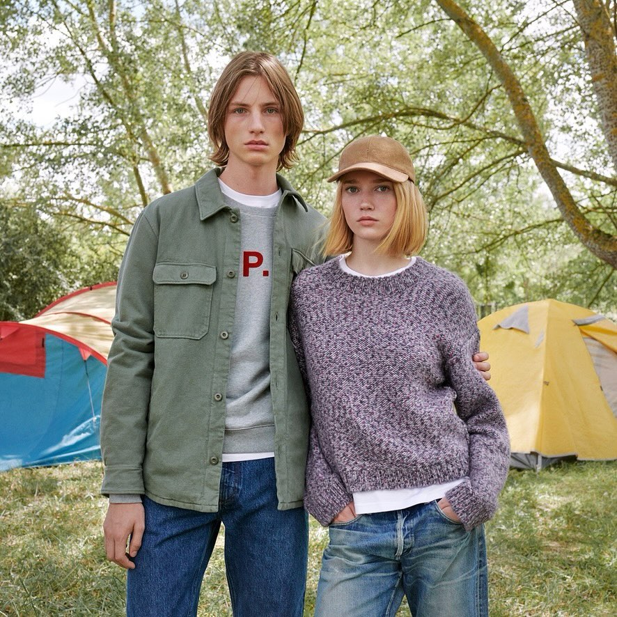 A young couple poses in a forest setting near tents. They are wearing casual clothing in a medium eye-level shot with natural lighting.