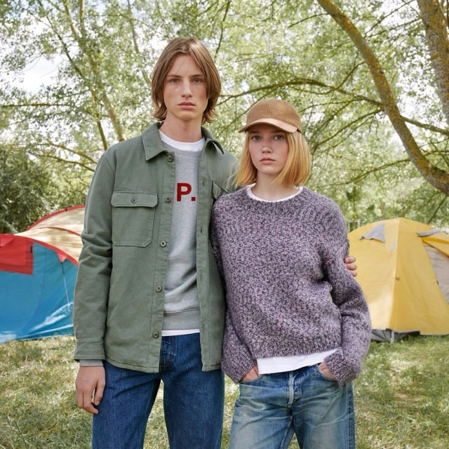 A young couple poses in a forest setting near tents. They are wearing casual clothing in a medium eye-level shot with natural lighting.