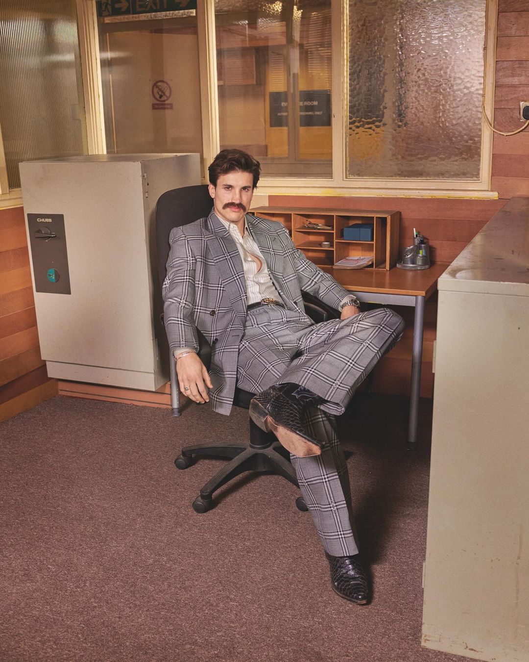 Adult man in a grey plaid suit sits at a desk with a computer and landline phone in an office setting with tan walls.