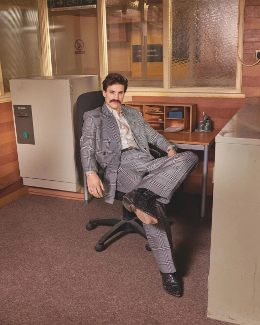 Adult man in a grey plaid suit sits at a desk with a computer and landline phone in an office setting with tan walls.