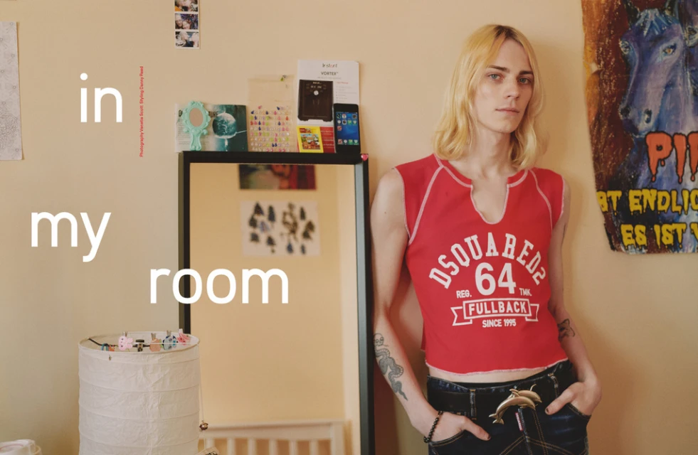 Young adult poses indoors wearing a red crop top and dark jeans in a casual indoor room setting with mirror and wall decor.