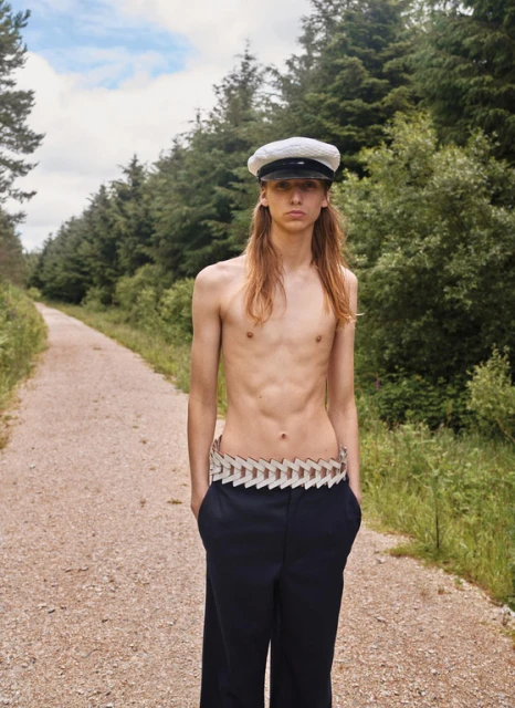 Shirtless person in sailor cap and trousers stands on dirt path, framed by trees and greenery on a partly cloudy day.