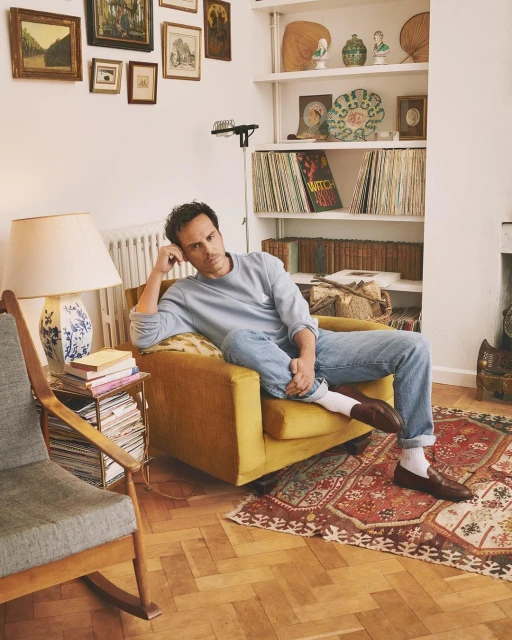 Man relaxes in a yellow armchair in a living room, bathed in warm, inviting light, surrounded by books, decor, and vintage charm.