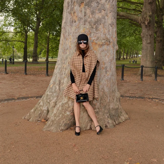 Woman in tan and black plaid cape poses against a large tree in a green park with handbag, black hat and sunglasses.