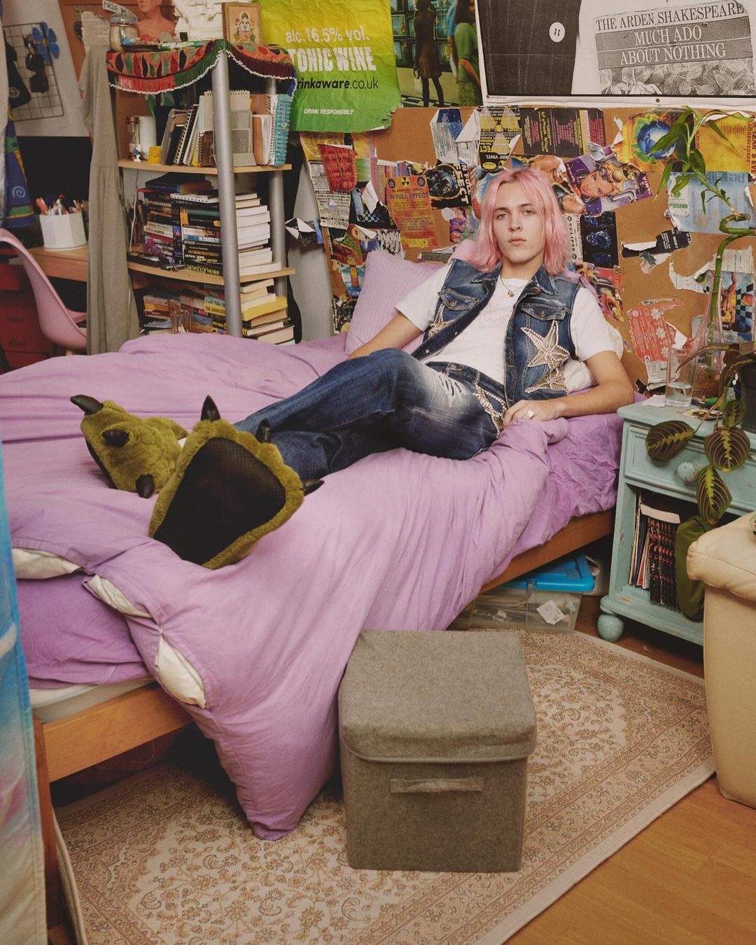Young adult with pink hair lounges on a lilac bed in bedroom with posters, wearing jeans and a denim vest with novelty monster slippers.