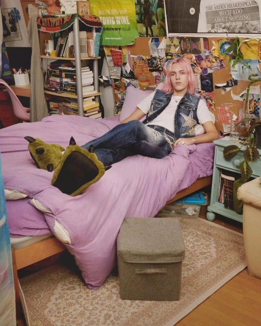 Young adult with pink hair lounges on a lilac bed in bedroom with posters, wearing jeans and a denim vest with novelty monster slippers.