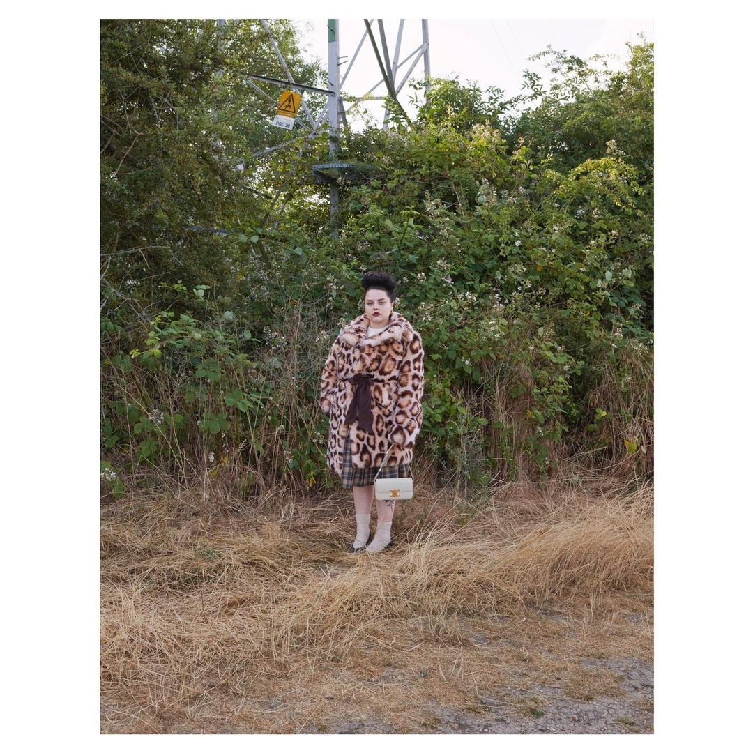 Adult female in an animal-print coat with a small purse, standing in a grassy, outdoor environment with an electrical tower in the background.