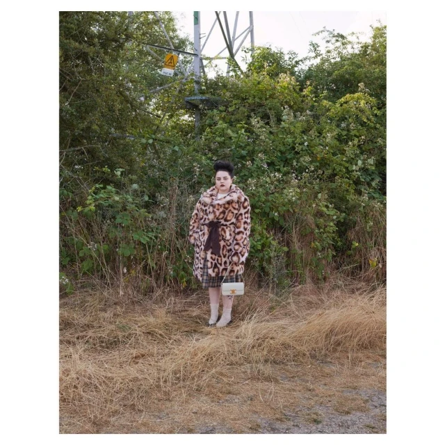 Adult female in an animal-print coat with a small purse, standing in a grassy, outdoor environment with an electrical tower in the background.