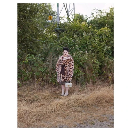 Adult female in an animal-print coat with a small purse, standing in a grassy, outdoor environment with an electrical tower in the background.