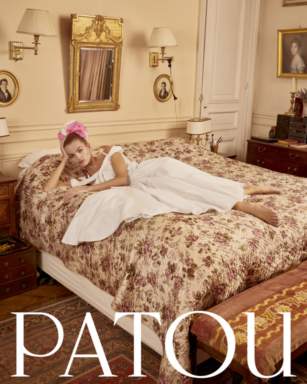 Woman in a white dress poses on a floral bed in a stylish, light-filled room with antique decor, creating an editorial fashion image.