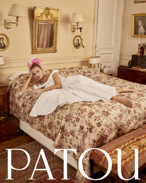 Woman in a white dress poses on a floral bed in a stylish, light-filled room with antique decor, creating an editorial fashion image.