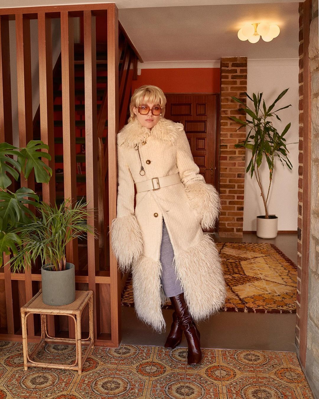 Woman in stylish vintage-inspired winter coat and brown knee-high boots poses indoors next to houseplants and wood-slatted wall.