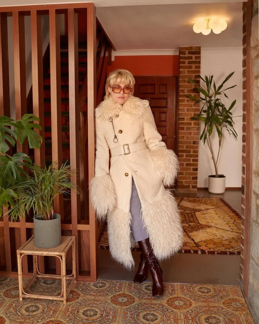 Woman in stylish vintage-inspired winter coat and brown knee-high boots poses indoors next to houseplants and wood-slatted wall.