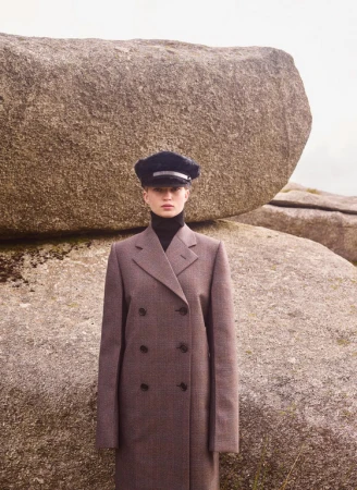 Model wearing a double-breasted brown coat and dark cap posed against large weathered rock formations.