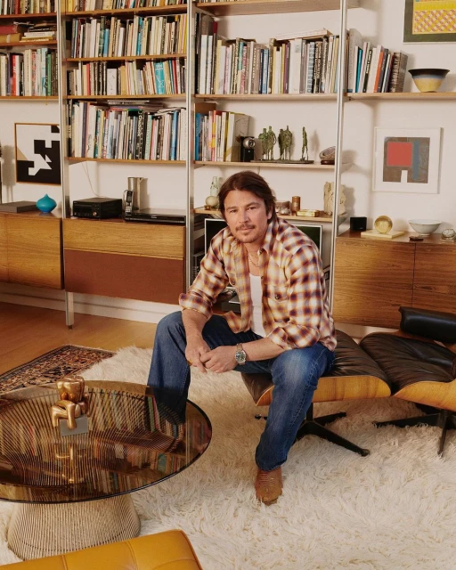 Adult male with mustache sits in black mid-century modern chair indoors. He wears jeans and a flannel shirt in a bright living room.