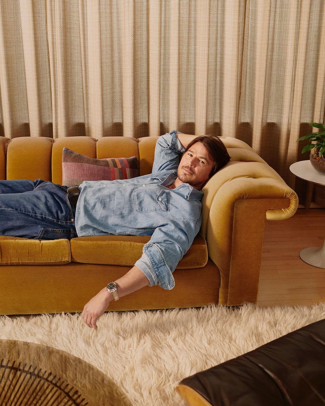 Adult male lounging on a yellow couch with a pillow, wearing denim and a watch in an indoor setting with neutral tones and shag rug.