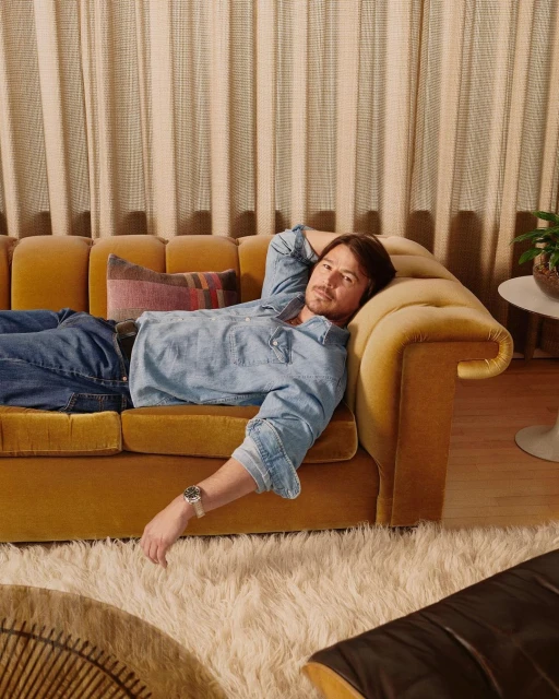 Adult male lounging on a yellow couch with a pillow, wearing denim and a watch in an indoor setting with neutral tones and shag rug.