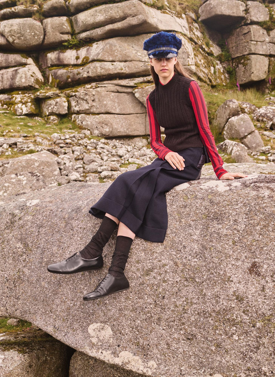 A young woman in a black vest, navy skirt, and blue cap sits on a rock formation outdoors, showcasing fashion against a natural backdrop.