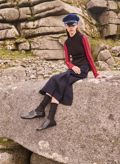 A young woman in a black vest, navy skirt, and blue cap sits on a rock formation outdoors, showcasing fashion against a natural backdrop.
