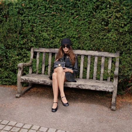 Woman in dark cap, sunglasses, and black leather coat sitting on a weathered wooden park bench holding a handbag