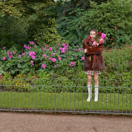 A woman in a fur coat stands behind a metal fence in a lush garden holding a bouquet of pink flowers.