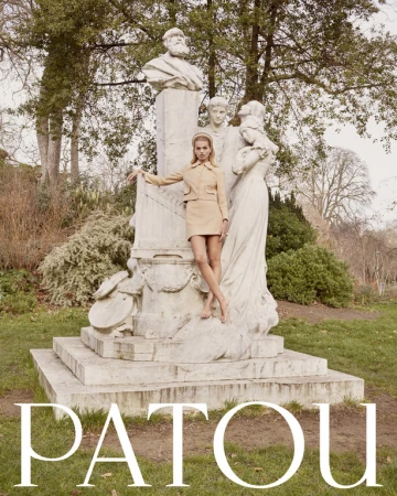 A fair-skinned woman in a beige suit poses by a white marble statue in an outdoor park setting with green grass and trees.
