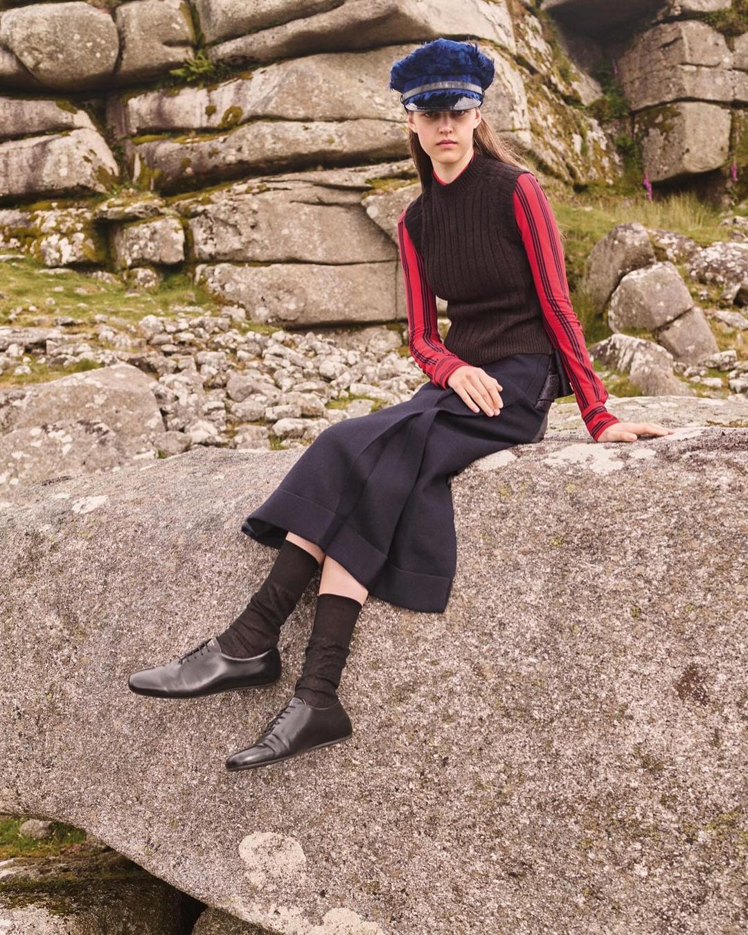 A young woman in a dark knit top, wide pants, and a cap sits on a large rock with a rocky landscape background in a fashion editorial.